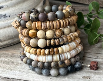 May include: A stack of eight bracelets made of various natural stones, including brown, beige, and grey. The bracelets are stacked on a wooden surface.
