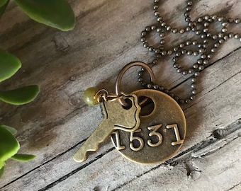 May include: A gold necklace featuring a key charm and a round tag inscribed with the number 1531. The necklace is displayed on a wooden surface with green foliage.