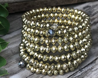 May include: A gold-toned bracelet made of faceted beads. The bracelet is wrapped around itself multiple times and has a small, dark grey bead at each end.