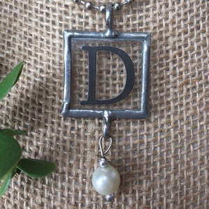 May include: A silver chain necklace with a square pendant featuring the letter "D" in black. The pendant has a small pearl charm hanging from it.