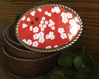 May include: A brown leather belt with a silver oval buckle featuring a red background with a white floral pattern.