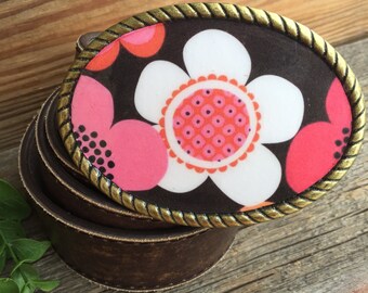 May include: A brown leather belt buckle with a floral design. The buckle features a large white flower with a pink centre and pink and orange petals. The background is black.