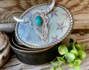 May include: A silver belt buckle featuring a turquoise stone and a stylised bull skull design. The buckle is set against a white background with stars.