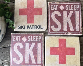 May include: Four square coasters with a ski theme. Two coasters feature a red cross with the text "Ski Patrol" in black. The other two coasters feature the text "Eat Sleep Ski" in white on a red background.