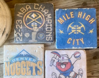 May include: Four square ceramic coasters featuring Denver Nuggets imagery. One coaster features the team's logo and the words "Denver Nuggets". Another coaster features the team's mascot, the Rocky Mountain  mascot, with the words "Denver Nuggets". A third coaster features the team's logo and the words "Mile High City". The final coaster features the team's logo and the words "22-23 NBA Champions".