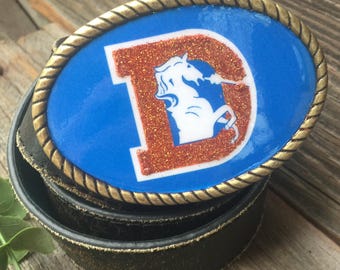 May include: A gold-toned belt buckle with a blue background and a white and orange glitter letter "D" with a white unicorn inside the letter.
