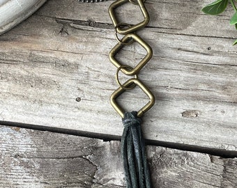 May include: A long necklace with a chain made of small metal beads. The necklace has three connected square-shaped metal charms and a long black tassel.