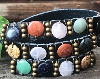 May include: A black leather wrap bracelet with multiple rows of colourful gemstone beads. The bracelet features round, flat beads in shades of green, pink, brown, and blue, interspersed with gold-toned beads.