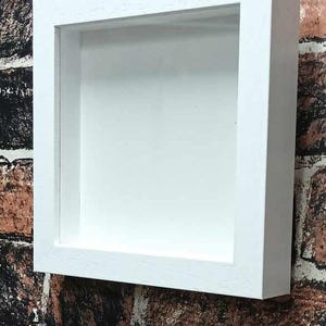 May include: A white, square shadow box frame with a recessed interior. The frame is mounted on a brick wall. The frame's interior is painted white, creating a clean, modern aesthetic. The frame is designed to display small objects.