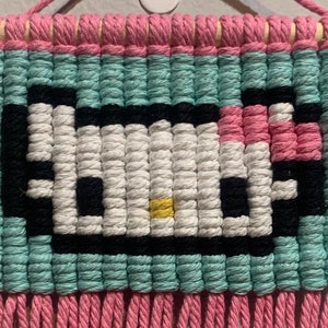 Może przedstawiać: Wiszący gobelin w kolorze różowym i turkusowym z pikselowym wzorem Hello Kitty. Gobelin ma różowe frędzle.