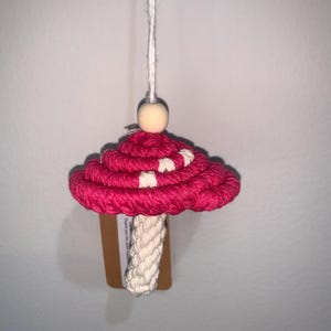 Puede incluir: Un adorno de hongo de crochet rosa y blanco con una cuenta de madera en la parte superior y una cuerda blanca para colgar.