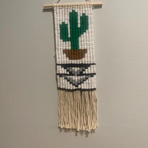 Pode incluir: Uma tapeçaria de parede em macramé com um cacto verde num vaso castanho sobre um fundo branco. O design inclui padrões geométricos pretos, cinzentos e brancos, com franjas bege na parte inferior. Uma cavilha de madeira e um cordão para pendurar estão no topo.