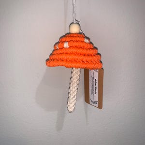 Puede incluir: Adorno de champiñón tejido en color naranja y blanco con una cuenta de madera en la parte superior. El adorno está colgado de una cuerda blanca y tiene una etiqueta marrón que dice "Velvet Rootz".