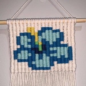 May include: A macrame wall hanging featuring a pixelated blue flower design with yellow and green accents. The artwork is crafted with off-white yarn and hangs from a wooden dowel, with fringe at the bottom.