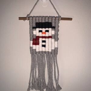 May include: A gray and white yarn wall hanging with a snowman design. The snowman has a black hat, orange nose, and red scarf. The hanging has gray tassels.
