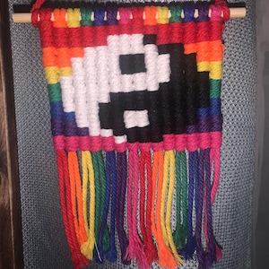 Op de afbeelding: Een handgemaakte macramé wandhanger met een gepixeld yin en yang symbool in zwart en wit. De bovenkant is een regenboog van kleuren, met een houten deuvel om op te hangen. De onderkant heeft kleurrijke franjes in regenboogtinten.