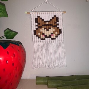 May include: A small, handmade macrame wall hanging featuring a pixelated fox design in shades of brown, tan, and pink against a white background. The wall hanging is suspended from a wooden dowel with a white cord and fringe.