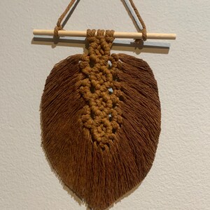 May include: A brown macrame wall hanging in the shape of a leaf. The leaf is suspended from a wooden dowel with a brown cord. The macrame has a detailed, textured center and fringed edges, creating a natural, bohemian aesthetic.