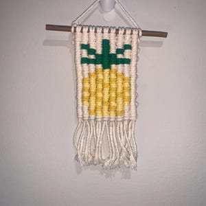 May include: A small woven wall hanging featuring a pineapple design. The pineapple is made with yellow and green yarn, and the background is white. The hanging has a wooden dowel at the top and white fringe at the bottom.