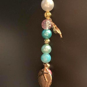 May include: A decorative hanging ornament featuring a variety of beads in shades of pink, teal, and white, interspersed with gold-colored seashell charms. A faceted crystal ball hangs at the bottom.