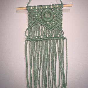 May include: Sage green macrame wall hanging. The handmade piece features a woven square design with a central circular element and long, dangling fringe. It is suspended from a natural wooden dowel and a matching cord for hanging. A decorative, bohemian-style wall art.