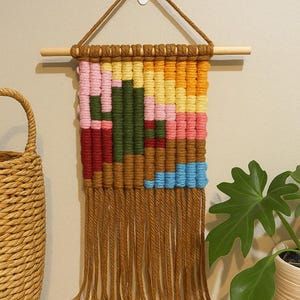 May include: A colorful macrame wall hanging with a geometric design. The piece features a wooden dowel and hanging cord. The woven design incorporates shades of brown, pink, green, yellow, and blue, with long brown fringe.