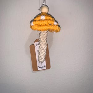 Puede incluir: Un adorno de hongo de crochet amarillo y blanco con una cuenta de madera en la parte superior. El hongo está colgado de una cuerda y tiene una etiqueta marrón que dice "Gracias por apoyar a las pequeñas empresas".