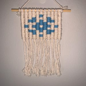 以下が含まれることがあります： クリーム色の紐に青と白の幾何学模様が織り込まれた小さな壁掛け。フリンジはクリーム色の紐でできています。