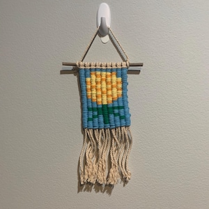 May include: A small wall hanging with a yellow flower design woven into a blue background. The hanging has a fringe of natural colored yarn.
