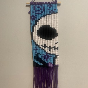 Può includere: Un arazzo da parete realizzato con filato in una combinazione di colori blu, bianco, nero e viola. Il filato è intrecciato a forma di teschio con una frangia sul fondo.