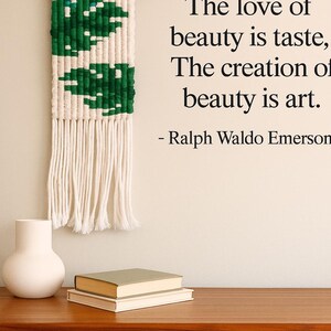 May include: A macrame wall hanging with a green leaf design hangs above a wooden cabinet. The text "The love of beauty is taste, The creation of beauty is art. - Ralph Waldo Emerson" is displayed on the wall. A white vase and two books are on the cabinet.