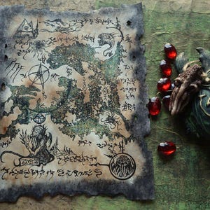 May include: A rectangular piece of aged, burnt-edge parchment featuring intricate black ink illustrations of a map, mythical creatures, and occult symbols, accompanied by several faceted red glass beads and a dark, textured sculptural object.