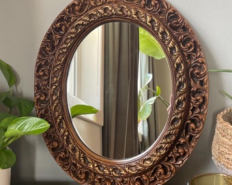May include: An oval-shaped mirror with a brown, ornate frame. The frame has a gold-colored design and is made of wood. The mirror is reflecting a room with a window and curtains.