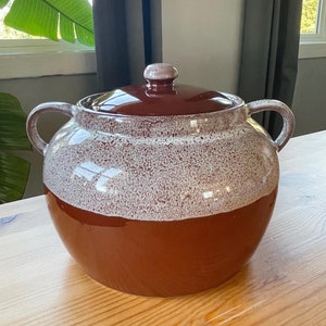 May include: A brown ceramic crock pot with a white glaze. The pot has two handles and a lid.
