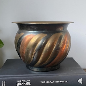 May include: A brass planter with a swirl design in shades of gold, copper, and black. The planter is sitting on top of two books.