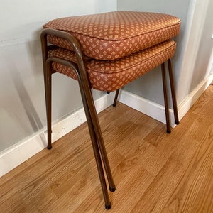 May include: Two brown upholstered stools with a geometric pattern, stacked on top of each other. The stools have a metal frame with four legs.
