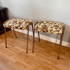 May include: Two vintage gold metal stools with floral patterned upholstery. The stools have a simple design with four legs and a rectangular seat.