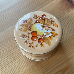 May include: A round, cream-colored ceramic trinket box with a floral design. The lid features a yellow apple, red berries, and brown leaves on a cream background.