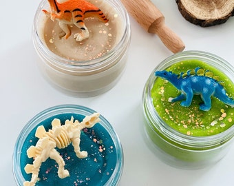 May include: Three clear containers of slime with dinosaur toys inside. The slime is green, blue, and beige. The dinosaurs are orange, blue, and white.