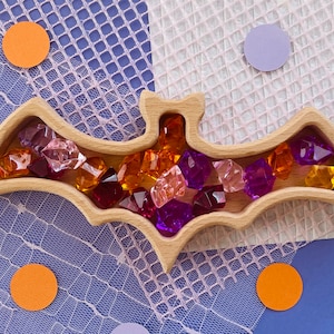 May include: A wooden bat-shaped dish with a decorative design, filled with colorful glass gems in shades of pink, purple, orange, and yellow.