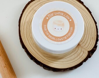 Puede incluir: Un recipiente blanco con una etiqueta redonda naranja que dice "*Marshmallow* Shop threeyellowstarfish.com" y un dibujo animado de malvavisco con una cara sonriente. El recipiente está sentado en una rebanada de madera.