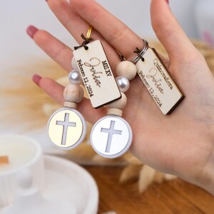 May include: Two wooden keychain charms with the name "Olivia" and the date "Febrero 12, 2024" engraved on them. The charms are attached to a string of wooden beads with silver and white beads. Each charm has a small round charm with a gold or silver cross.