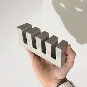 May include: A rectangular gray concrete object featuring four vertical slots cut into the top edge, creating a comb-like structure with five distinct teeth. The surface of the material displays a mottled texture with variations in light and dark gray tones.
