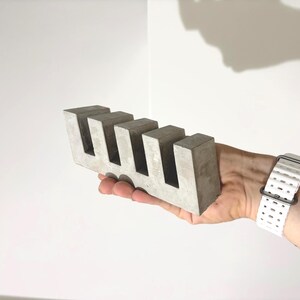 May include: A rectangular gray concrete block featuring four evenly spaced vertical cutouts along the top edge. The object has a smooth, matte finish and a solid, geometric construction with sharp, clean edges and a uniform light gray color throughout.