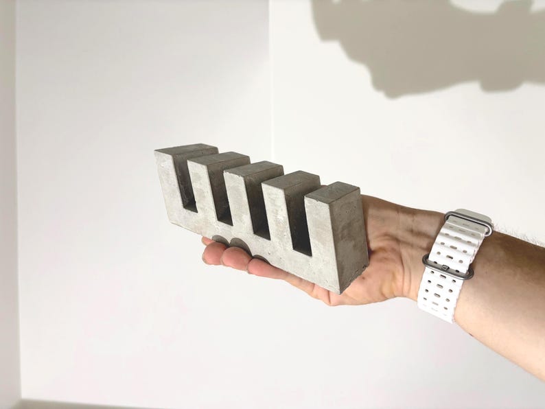 May include: A rectangular gray concrete block featuring four evenly spaced vertical cutouts along the top edge. The object has a smooth, matte finish and a solid, geometric construction with sharp, clean edges and a uniform light gray color throughout.