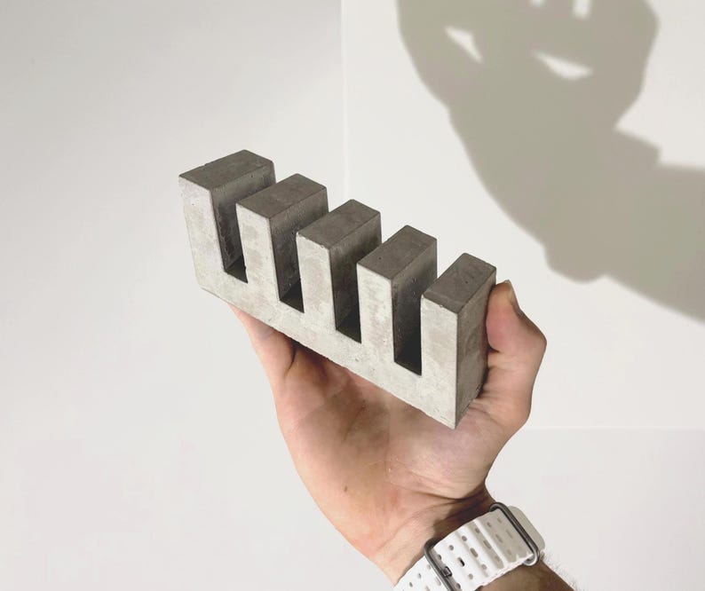 May include: A rectangular gray concrete object featuring four vertical slots cut into the top edge, creating a comb-like structure with five distinct teeth. The surface of the material displays a mottled texture with variations in light and dark gray tones.