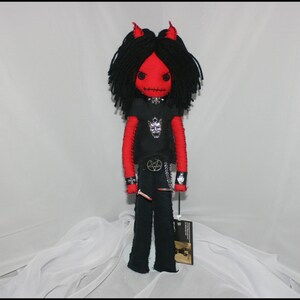 May include: A red and black felt doll with horns, wearing a black t-shirt with a silver pentagram design and black pants. The doll has black yarn hair and is holding a small red object in its hand.