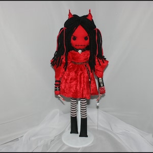 May include: A red velvet doll with black yarn hair and horns. The doll is wearing a red velvet dress with black and white striped stockings and black shoes. The doll is standing on a white pedestal.