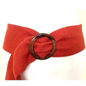 May include: A red linen belt with a large, brown, round buckle. The belt is tied in a knot at the front.