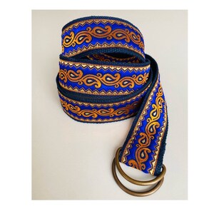 Puede incluir: Un cinturón azul y dorado con estampado y hebilla de metal. El cinturón está hecho de tejido tejido y tiene un diseño de paisley.